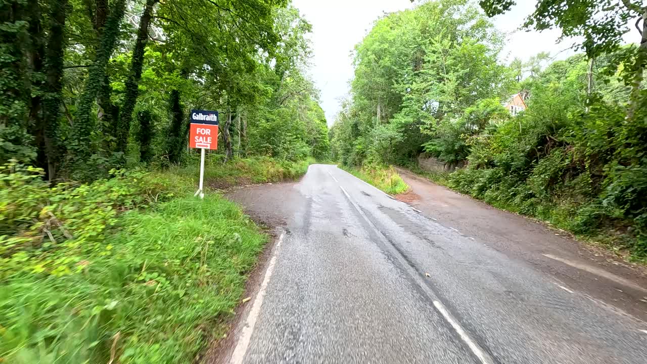 A vehicle travels down a winding, tree-lined rural road in Cromarty, Scotland, under overcast daylight. The camera moves smoothly forward, capturing lush greenery and roadside signs
