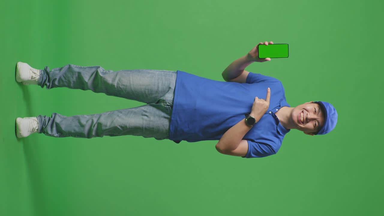 Full Body Of Asian Male Courier In Blue Uniform Smiling And Pointing To Green Screen Smartphone While Standing On Green Screen Background In The Studio