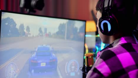 close up back view of asian boy streamer playing car racing game over network on computer, talking on the microphone.  live stream video game, desk illuminated by rgb led strip light