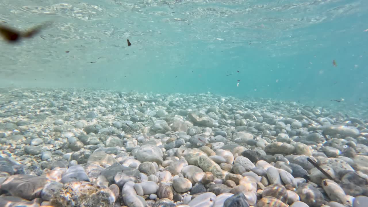 Underwater view of smooth round pebbles on seabed in crystal clear turquoise sea water with shimmering sunlight rays from surface