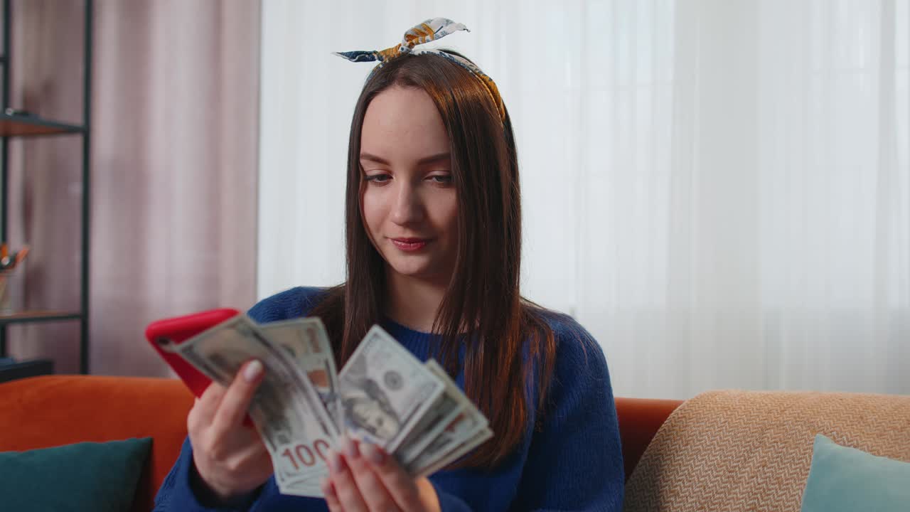 Smiling happy young woman holding cash money and mobile phone calculate domestic bills at home