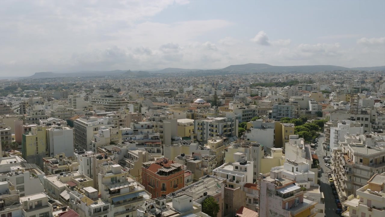 Vista aerea con drone sui tetti di Heraklion, Creta, che mostra la fitta espansione urbana, una chiesa a cupola e le colline che si ergono all'orizzonte sotto nuvole soffici.