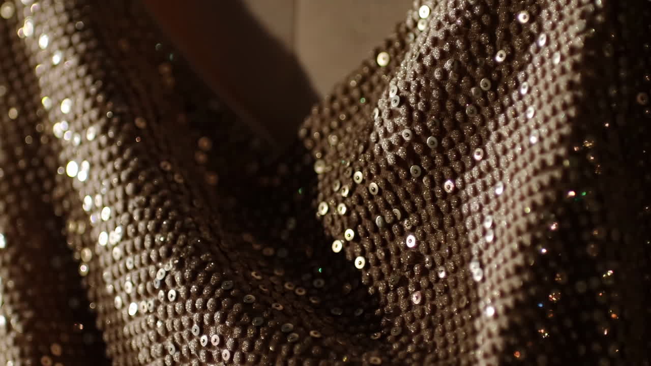 Tessuto di paillettes scintillante drappeggiato in una calda luce dorata, stilista di alta moda che leviga le pieghe, lenta panoramica cinematografica
