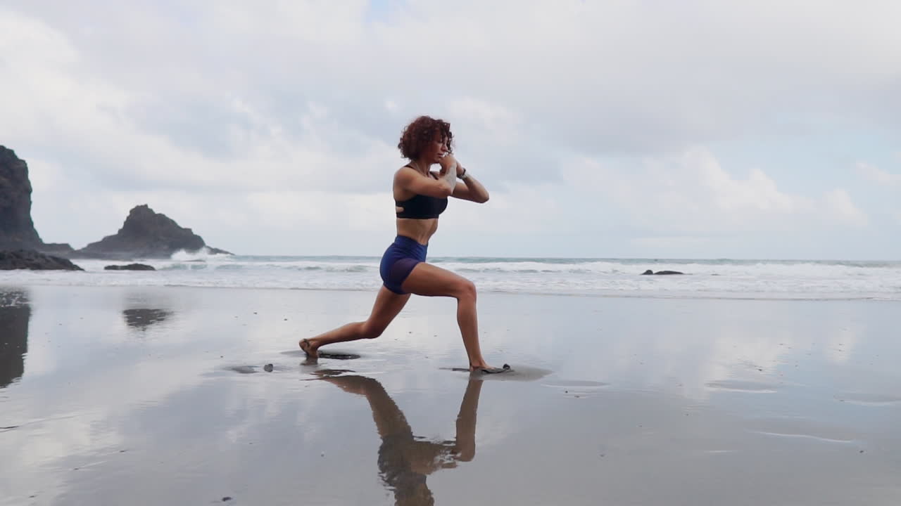 A young, slender woman engages in stretching and yoga by the ocean's edge, her gaze set on the horizon, all captured in slow motion