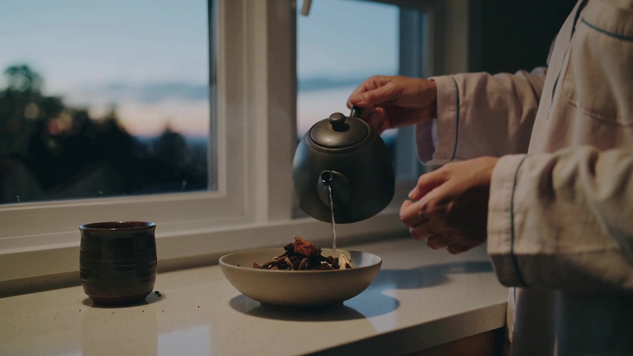 Pouring woman in pajama top tilting green teapot into bowl at dusk, preparing infusion, copy space