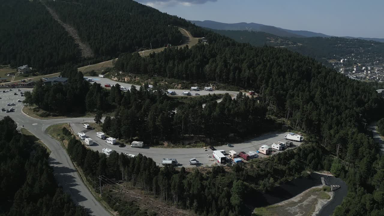 Filmato aereo dall'alto che cattura un parcheggio di montagna curvo pieno di camper, circondato da fitte foreste sempreverdi e dolci colline sotto un cielo limpido