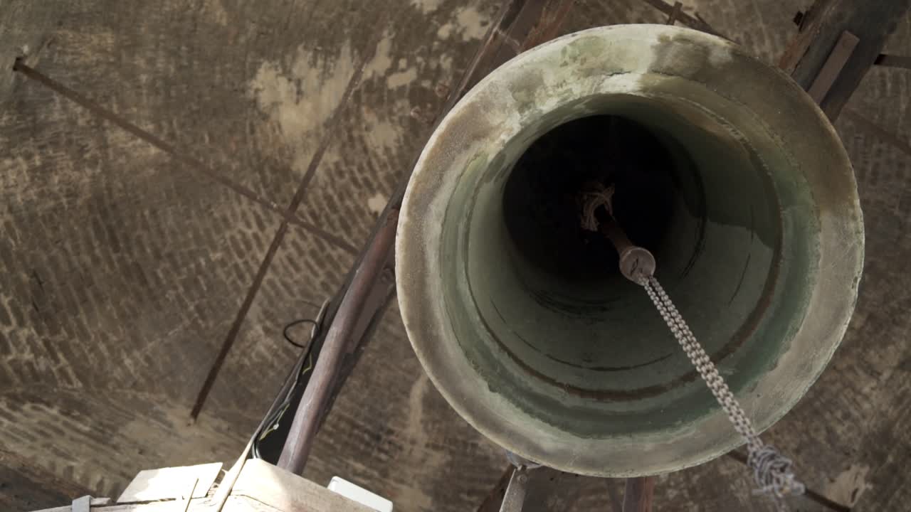 The large, aged, and rustic bell is fastened to the ceiling and held up by an iron beam
