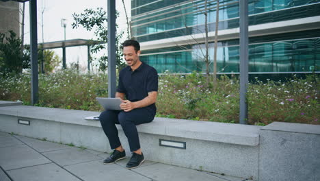 smiling businessman videocalling laptop at bus stop. freelancer man waving hand