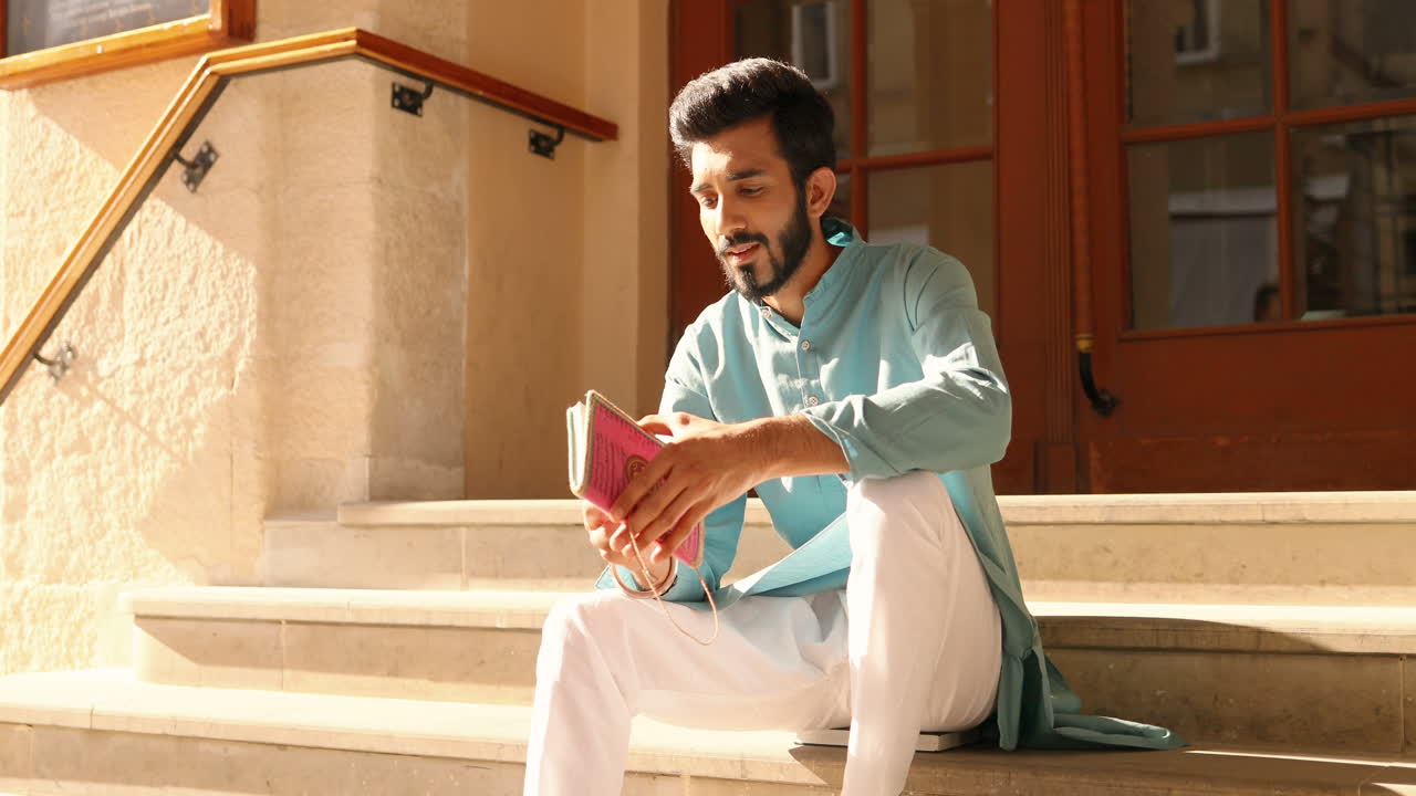 Handsome young Hindu man in traditional clothes sitting on steps outdoors and reading notes in notebook. Male from India checking planner. Day planning. Diary.