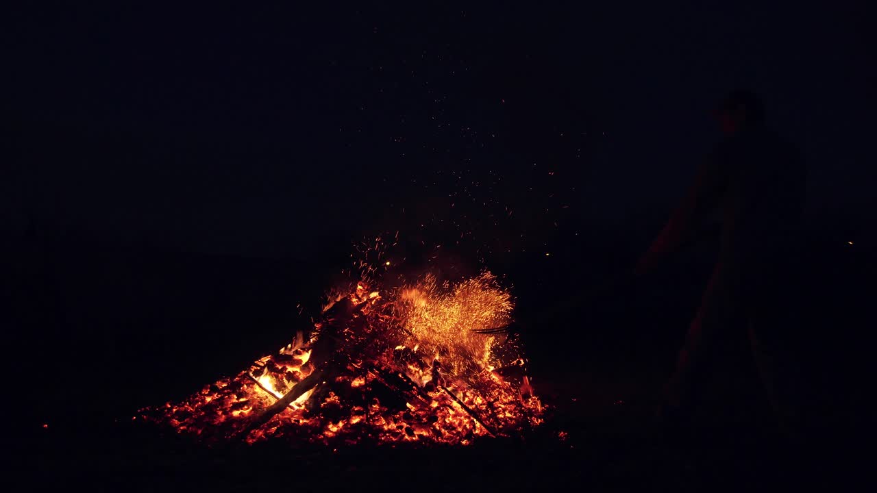 a man straightens forks burning coals in the fire. sparks of a fire against the dark sky