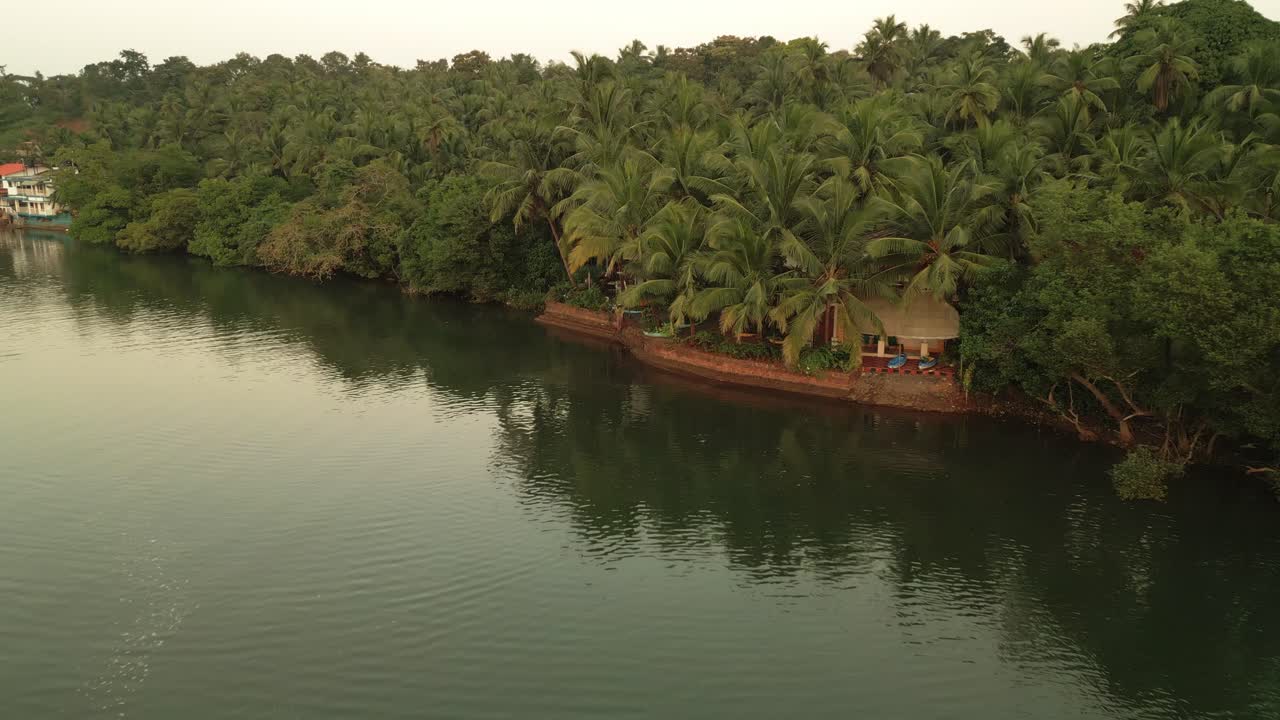 A drone view of a serene lake surrounded by lush green trees on both sides, with towering mountains in the background. The calm water perfectly reflects the vibrant greenery Goa India