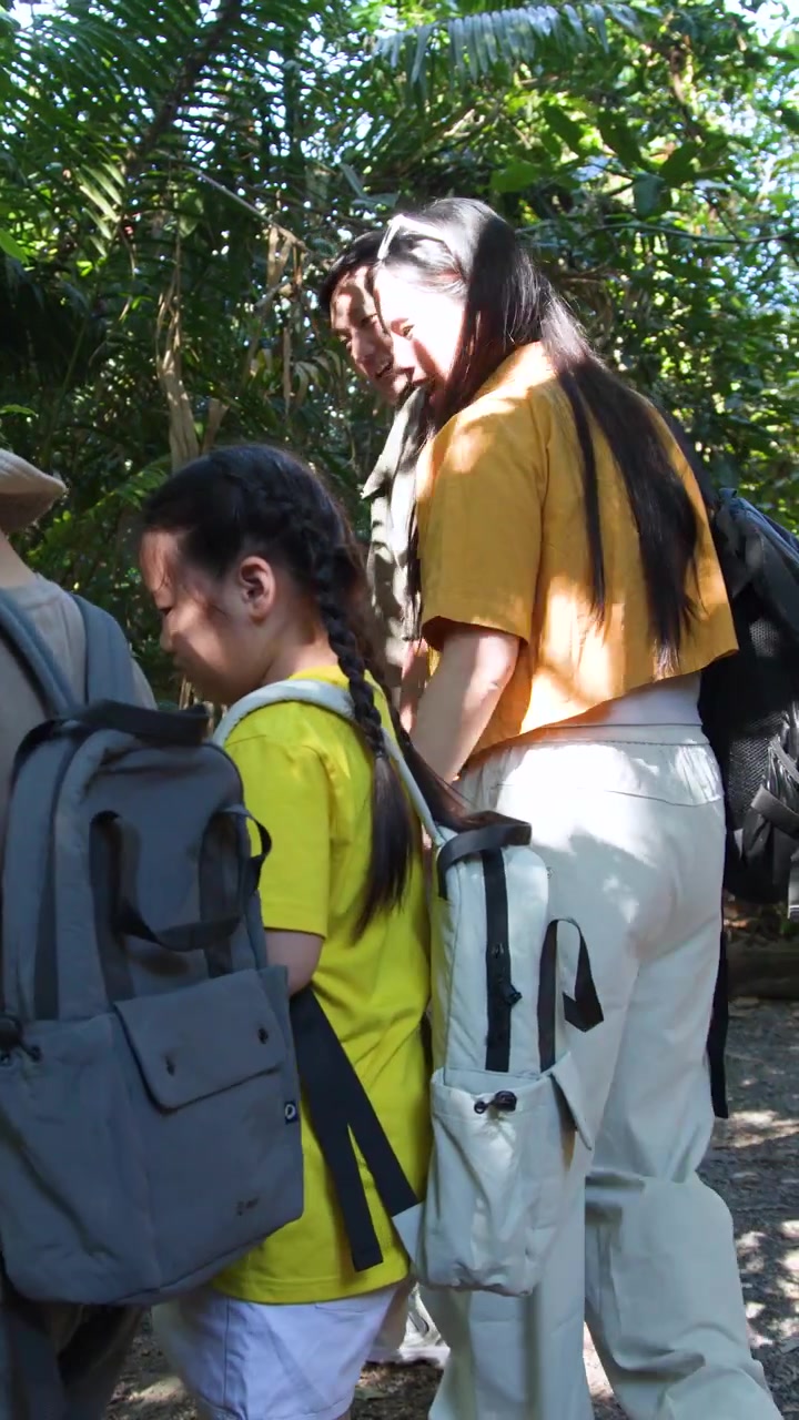 Thai family wearing backpacks explores a tropical forest trail under bright natural sunlight