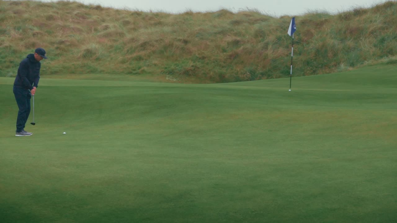 남자 골퍼는 부프 앤 런 을 사용하여 아일랜드 링크 골프 코스의 구을 향해 그린을 아내고, 비가 오는 동안 공을 향해 느린 동작으로 걸어갑니다.