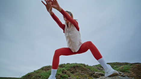 brunette practicing fitness exercises in red leggings at gloomy rocky hills