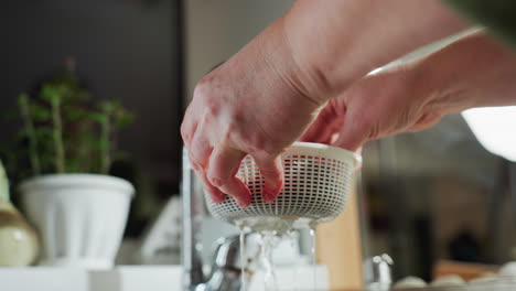 close up of hands holding white plastic basket filled with rinsed berries over sink as water drips through basket, emphasizing food hygiene, and domestic kitchen routine with natural lighting
