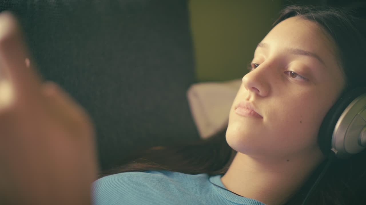 Teen girl relaxing at home, listening to music with headphones and using her smartphone. A calm, intimate moment of digital leisure in a cozy setting.
