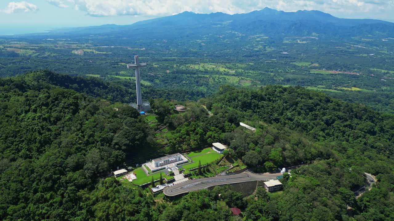 Left pan aerial showcasing the towering Mt. Samat National Shrine, revealing its towering cross monument framed by lush forested hills and sweeping valleys in Pilar, Bataan, Philippines