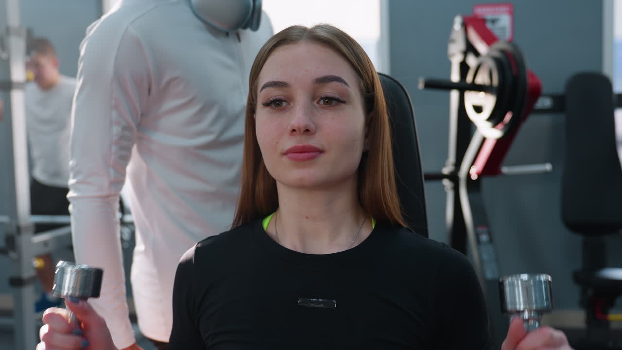 trainer in white shirt with neck headphones handing two metal dumbbells to female trainee seated in gym chair guiding form as outside window reveals moving car in background behind weight rack mirror