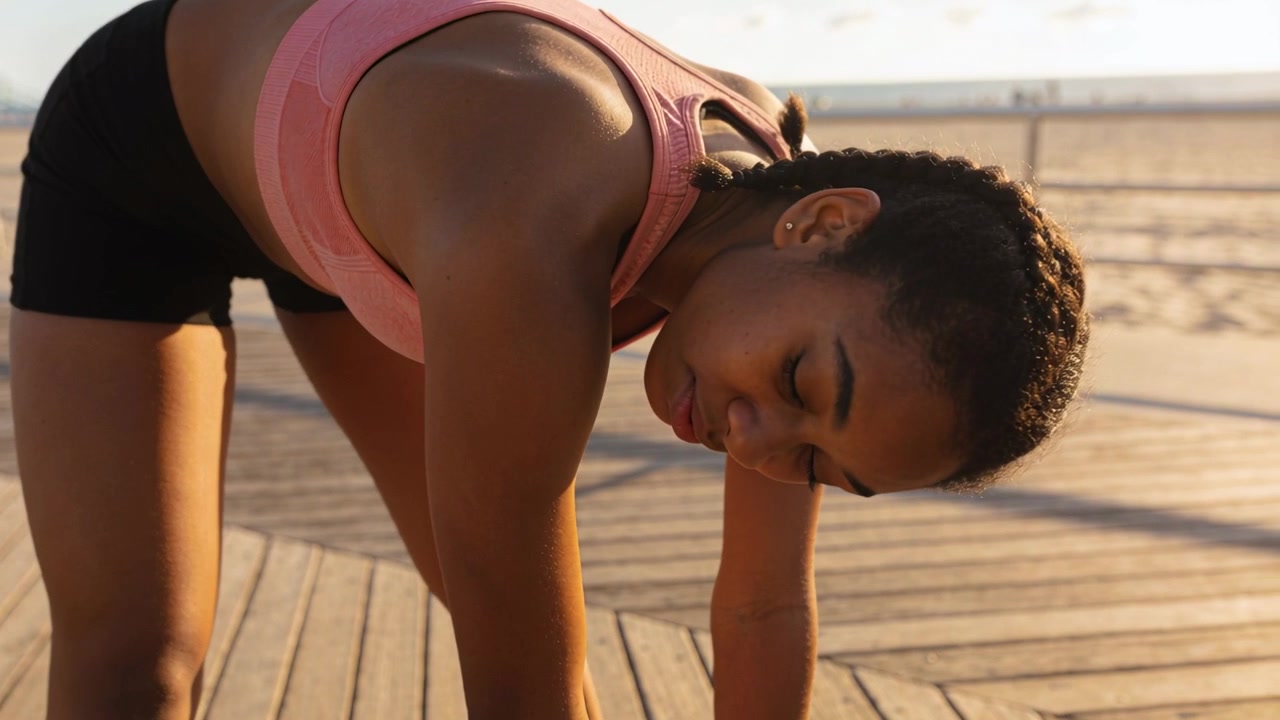 Stretching from forward fold, woman in pink sports bra lifting head, torso on boardwalk by ocean