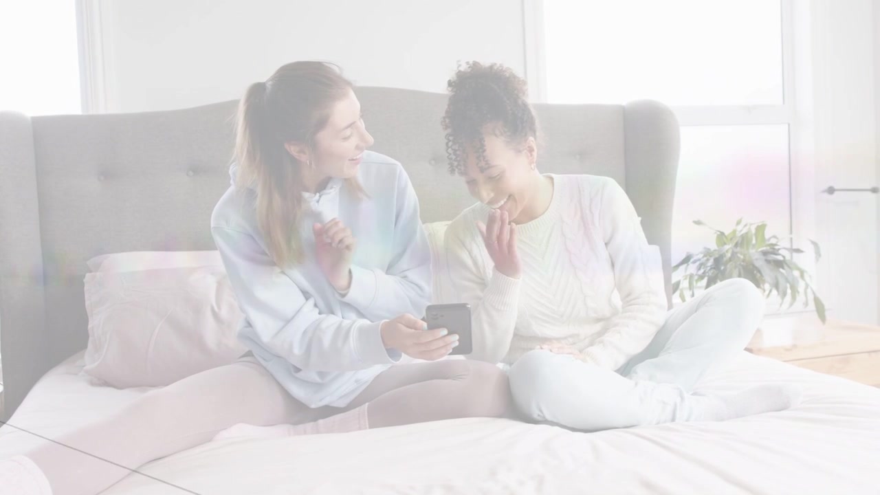 Left woman angling phone, right woman gasping laughing, exposure flicker, viewing tech clip on bed