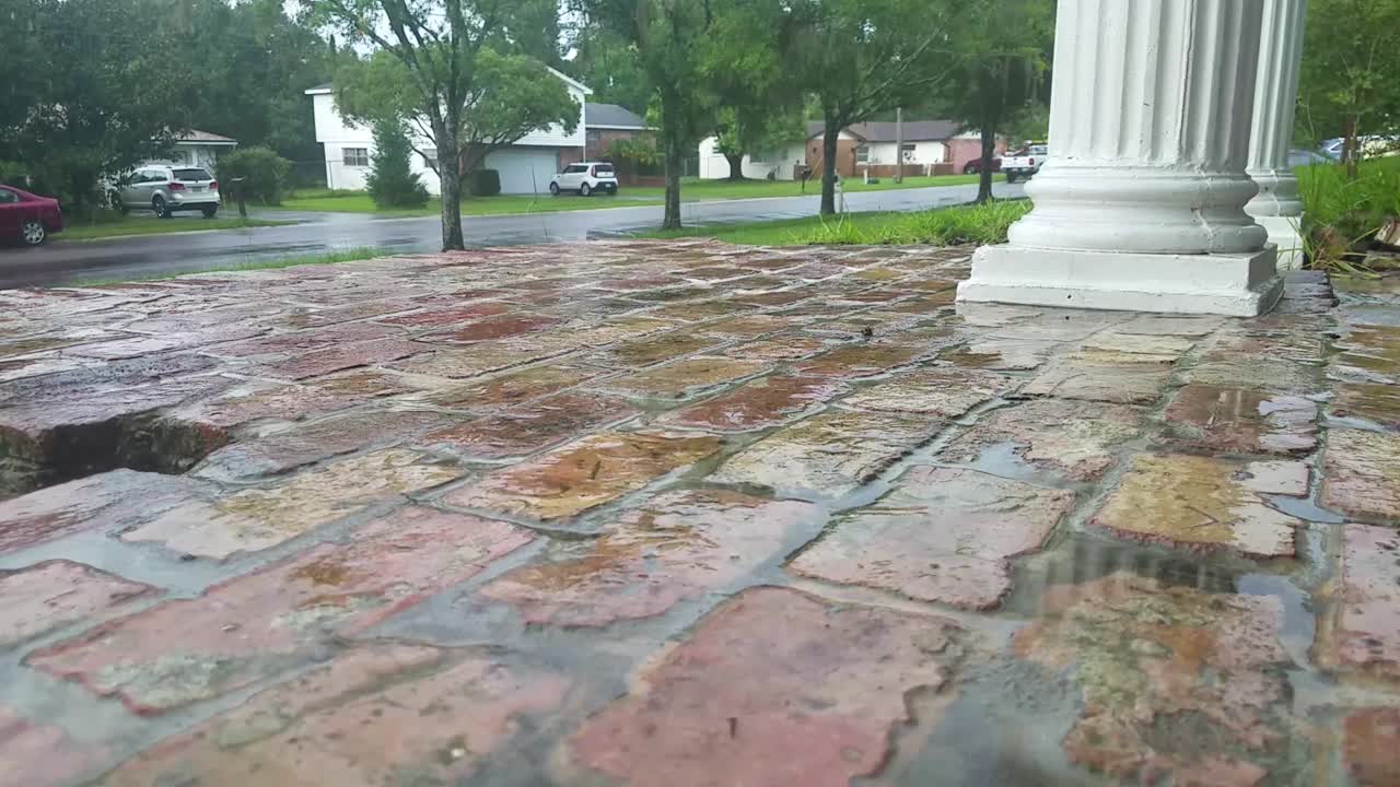 Rain drops on brick pavement, long shot, rainy weather, cars and houses in background.