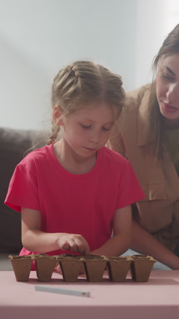 Little girl puts seeds into seedling tray with mother and younger brother in room slow motion. Woman teaches daughter to grow plants at family labor education