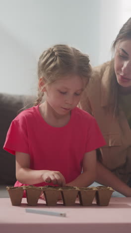 little girl puts seeds into seedling tray with mother and younger brother in room slow motion. woman teaches daughter to grow plants at family labor education