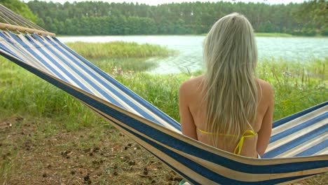 young beautiful blonde girl sitting and swinging on blue and white stripy hammock by the lake looking out and enjoying the view