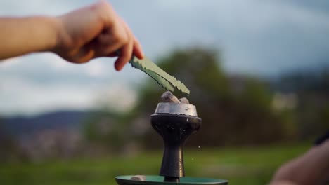 hand holding tongs places hot coal on a hookah outdoors during a relaxed sunny day