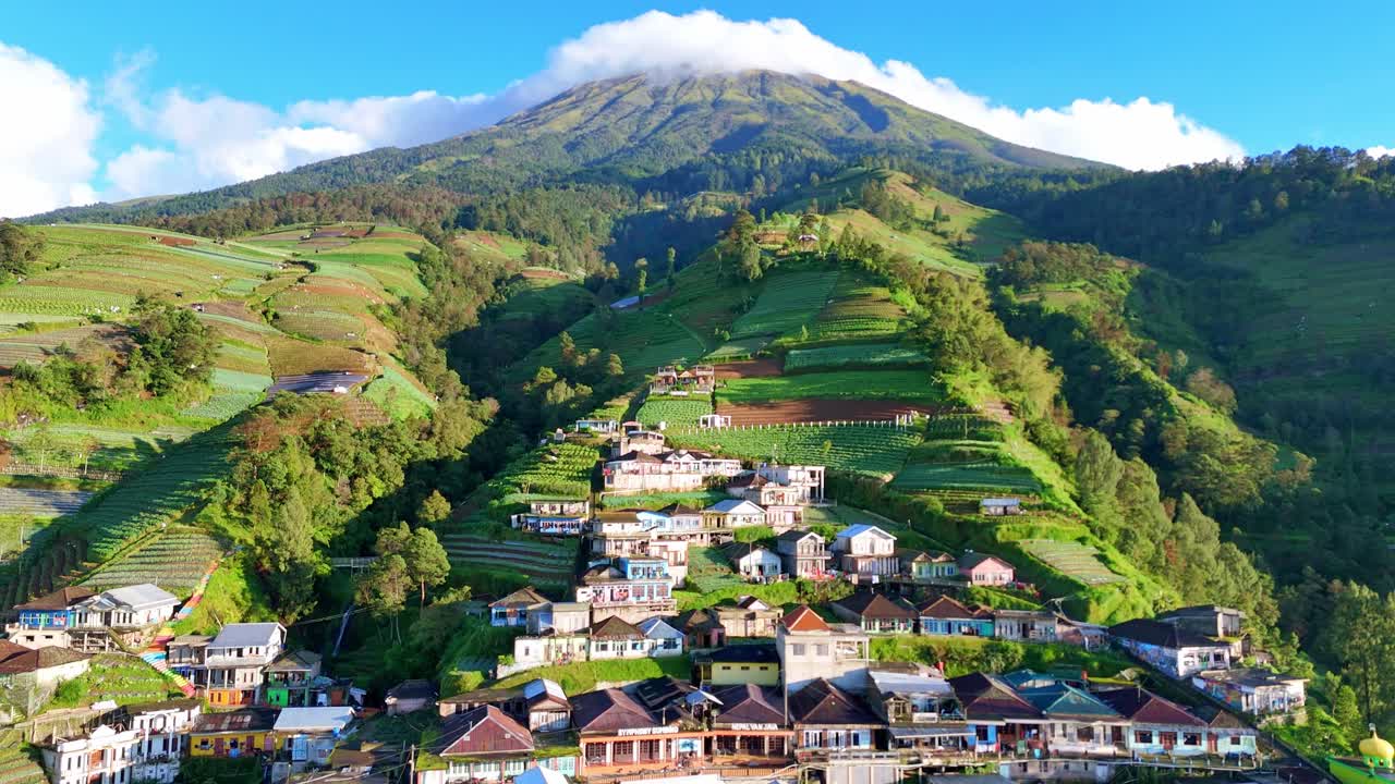Drone footage showcasing Nepal van Java, a picturesque mountain village on the green hills of Mount Sumbing. Vibrant homes, farmlands, and a mosque highlight the harmony of nature and culture