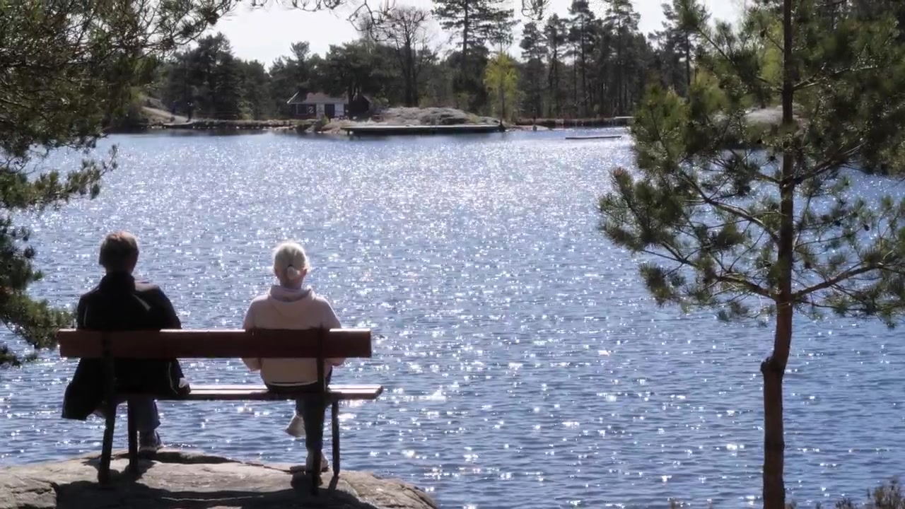 Coppia anziana che guarda un lago mentre si gode una giornata di sole. Ampia inquadratura nella natura su una panchina, a due piedi di distanza.