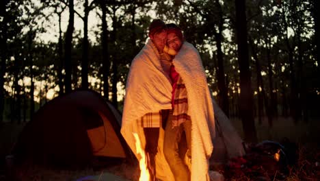 A-happy-couple,-a-guy-and-a-girl,-stand-near-an-evening-fire-wrapped-in-a-white-blanket-and-hug-and-communicate-with-each-other-against-the-backdrop-of-the-floor-and-the-evening-autumn-forest-during-a-hike