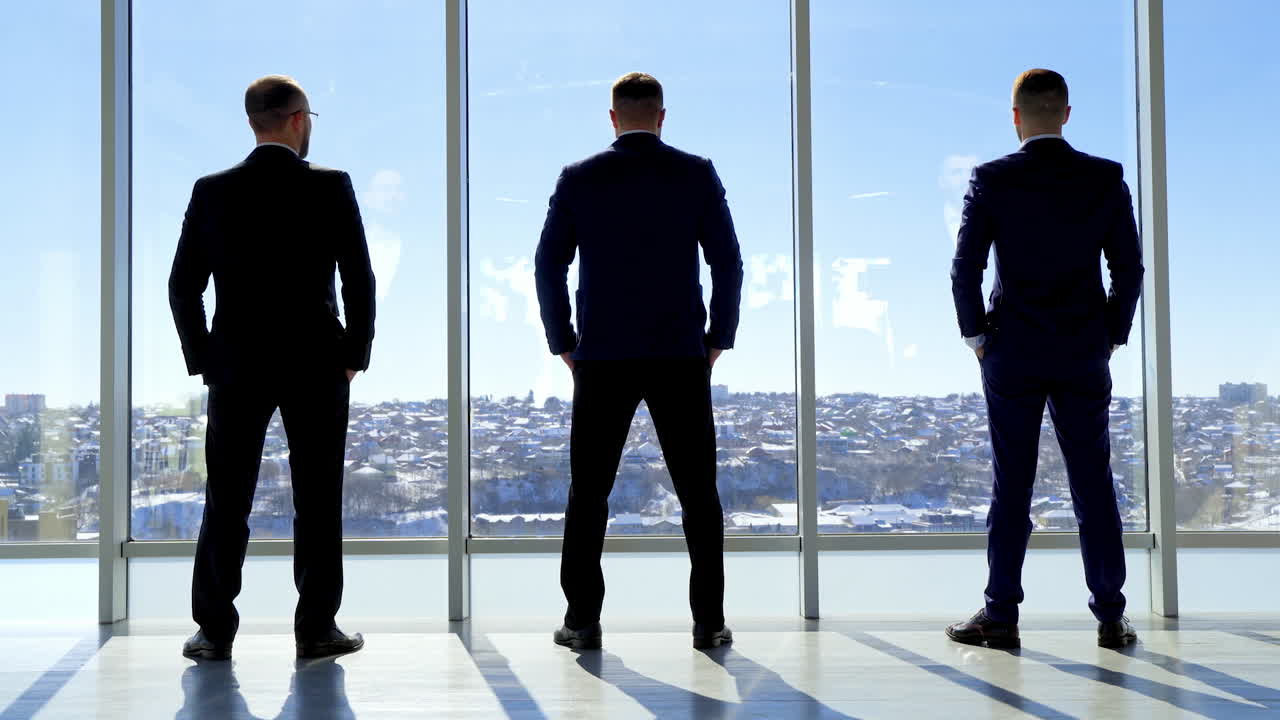 Businessmen looking out of large office window. Back full-length portraits of three entrepreneurs in costumes standing by the window in a office at daytime. Rear view.
