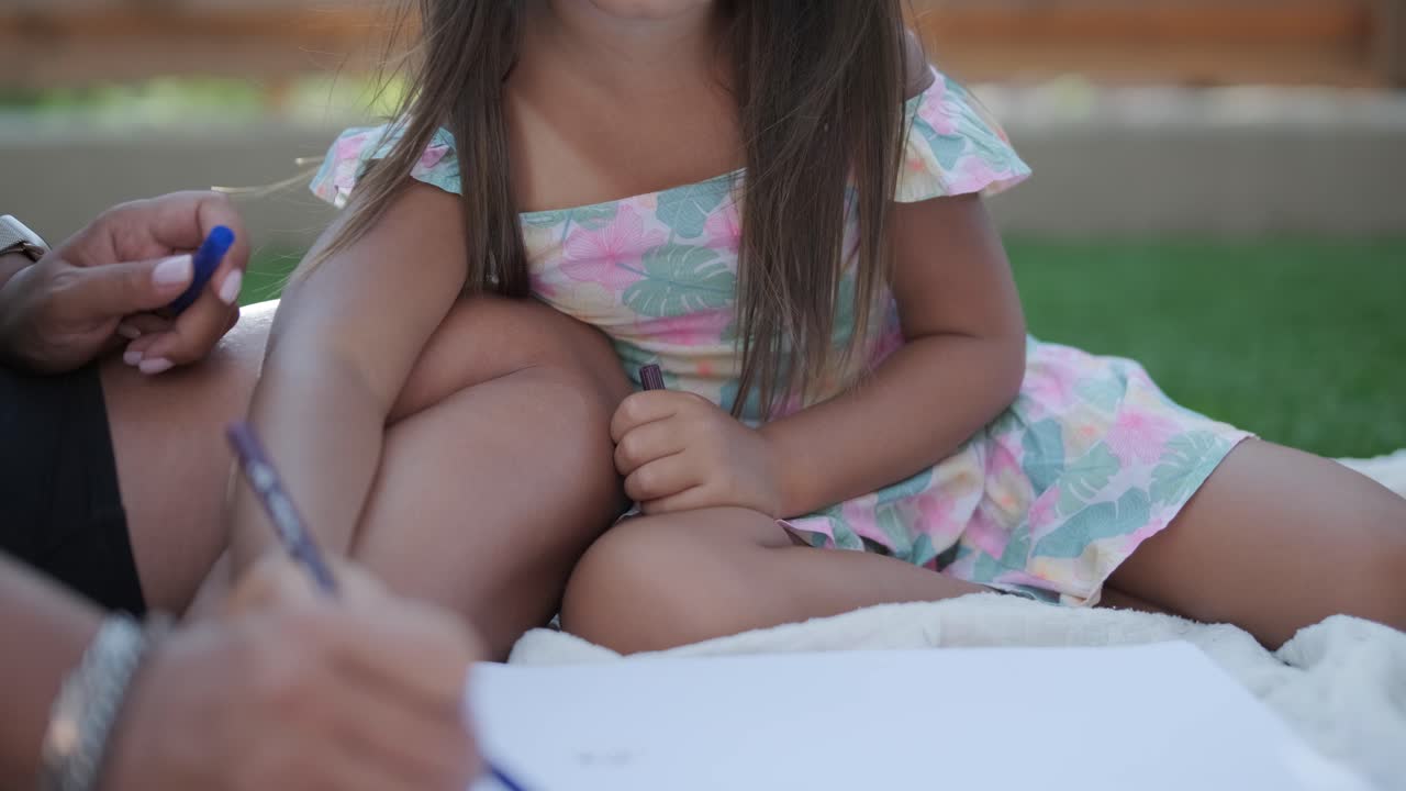 Una madre e la sua giovane figlia sono sedute su una coperta nel cortile, impegnate in un'attività di disegno insieme. La scena all'aperto cattura un momento familiare sereno, promuovendo la creatività e il legame