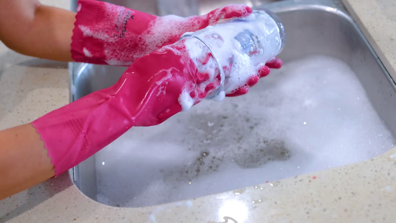 Hands in pink gloves wash a glass with a sponge in a kitchen sink, surrounded by soapy water