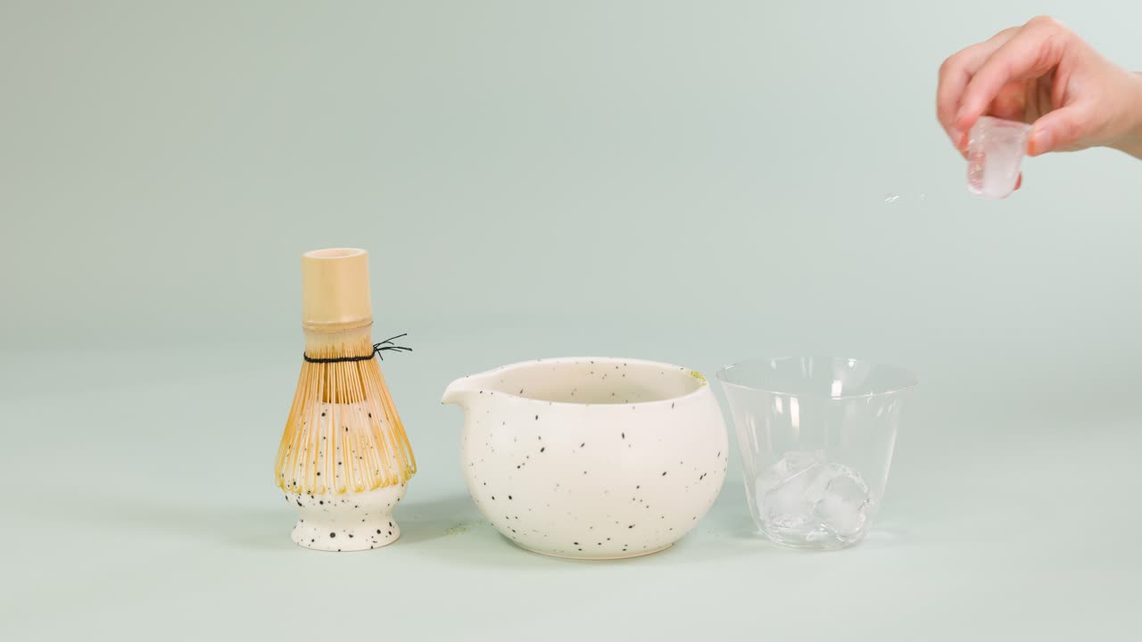 Hand placing ice cubes in glass beside ceramic bowl and bamboo whisk under soft lighting