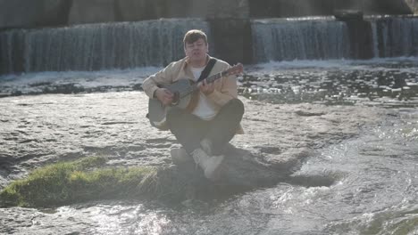 young man playing guitar sitting on the bank of a mountain river on a background of rocks. concept of freedom relaxation. place