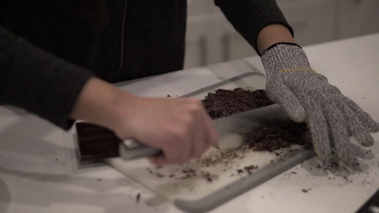 A young girl cuts up dark chocolate before melting it for millionaire shortbread cookies while wearing safety gloves-1