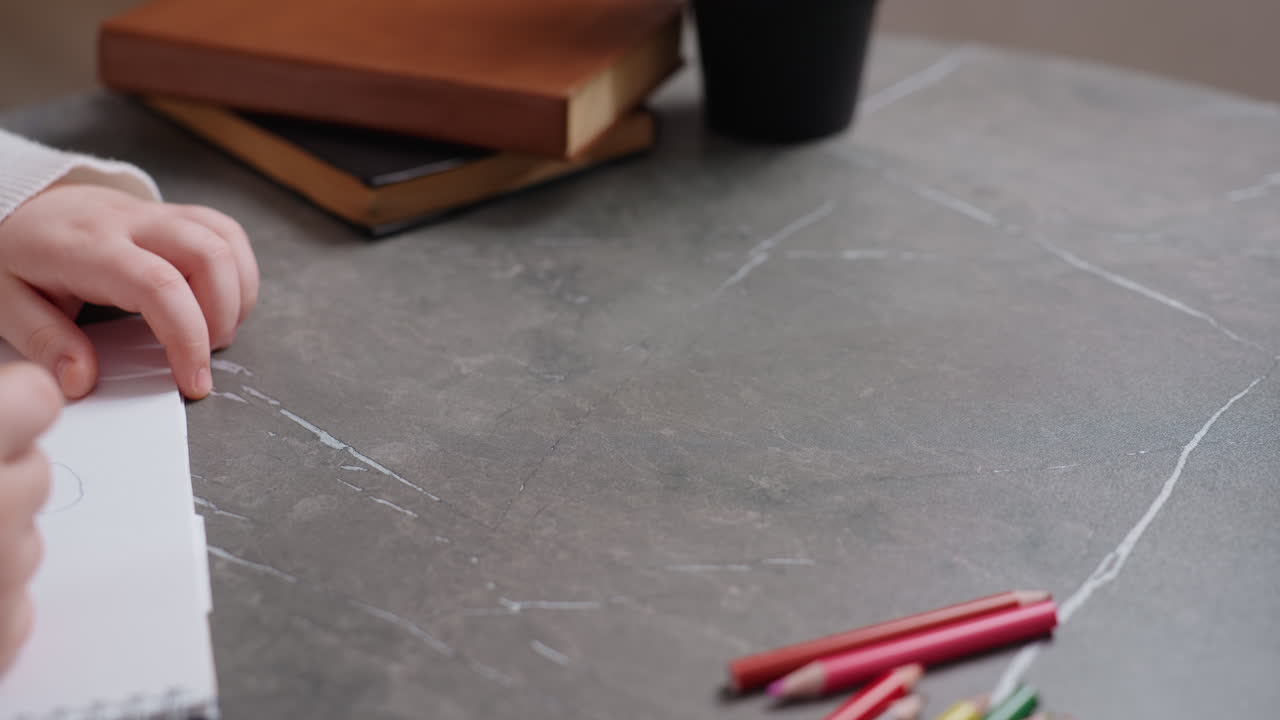 Close up of girl hand holding pencil while parent gestures during drawing activity at table with scattered colored pencils, showing interaction, guidance, creativity, education at home