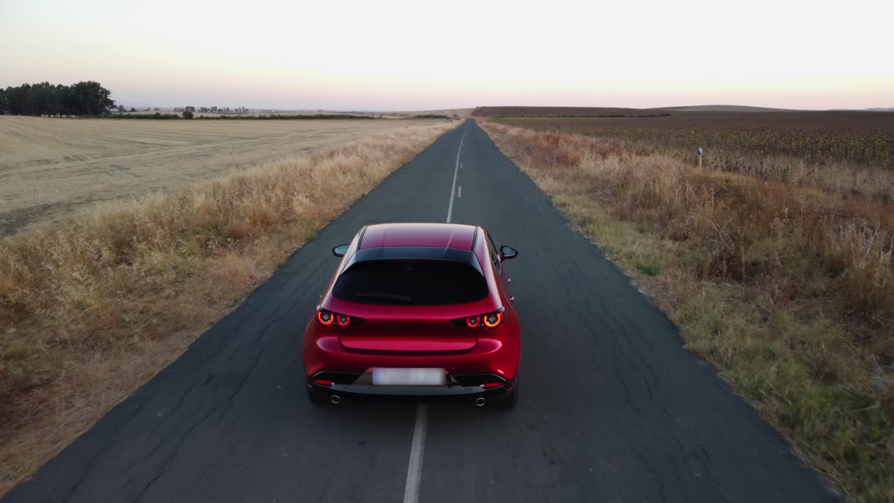 Shiny new red car drives slowly on narrow road through crop fields