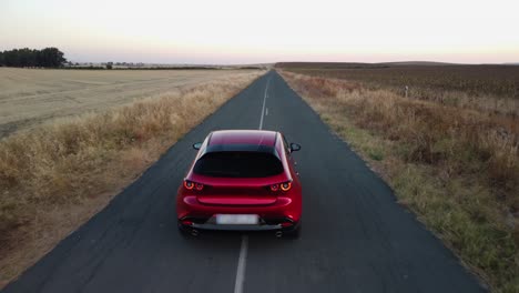 shiny new red car drives slowly on narrow road through crop fields