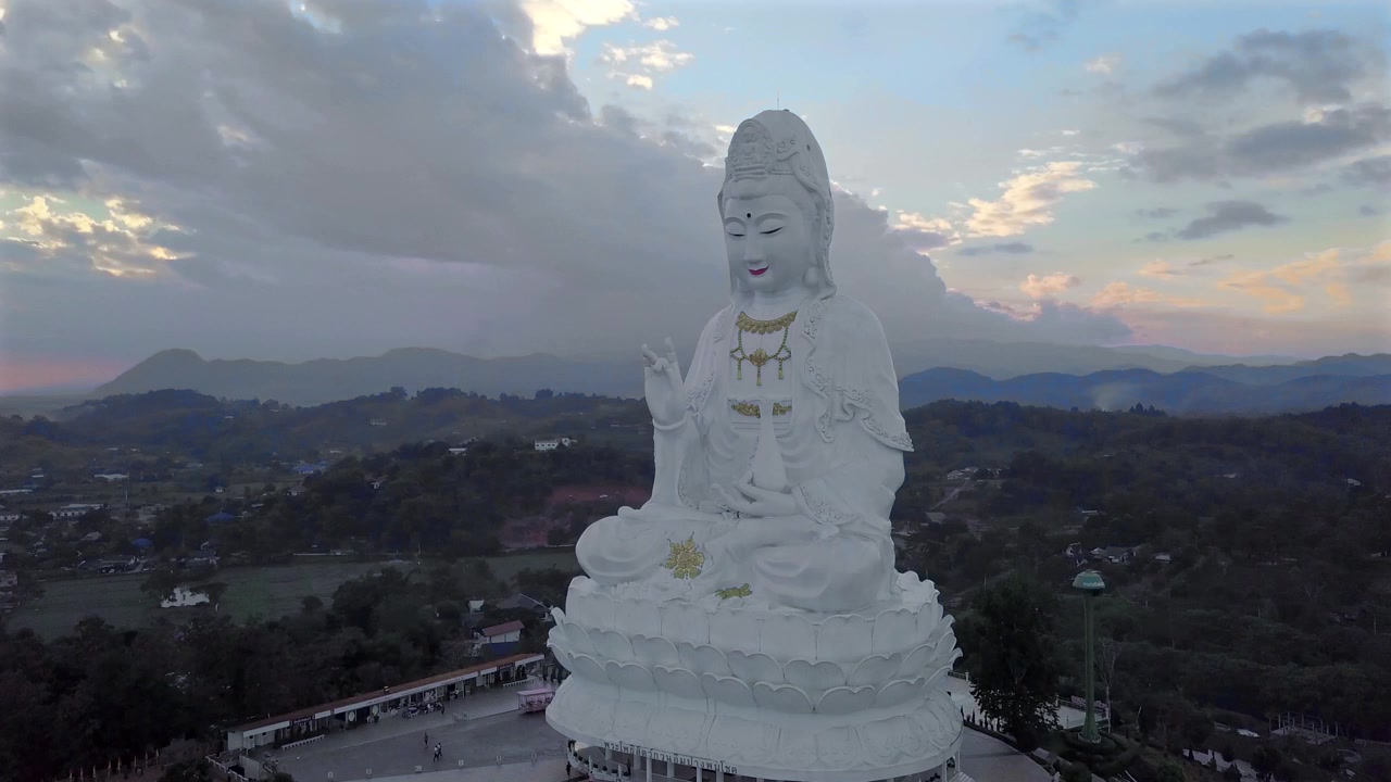 Thailand, Chiang Rai, Wat Huay Pla Kang. Droneoptagelse af Buddha-statuen, der drejer horisontalt rundt om den hvide statue fra venstre mod højre, med hvide og lyserøde skyer på den blå himmel under solnedgang.