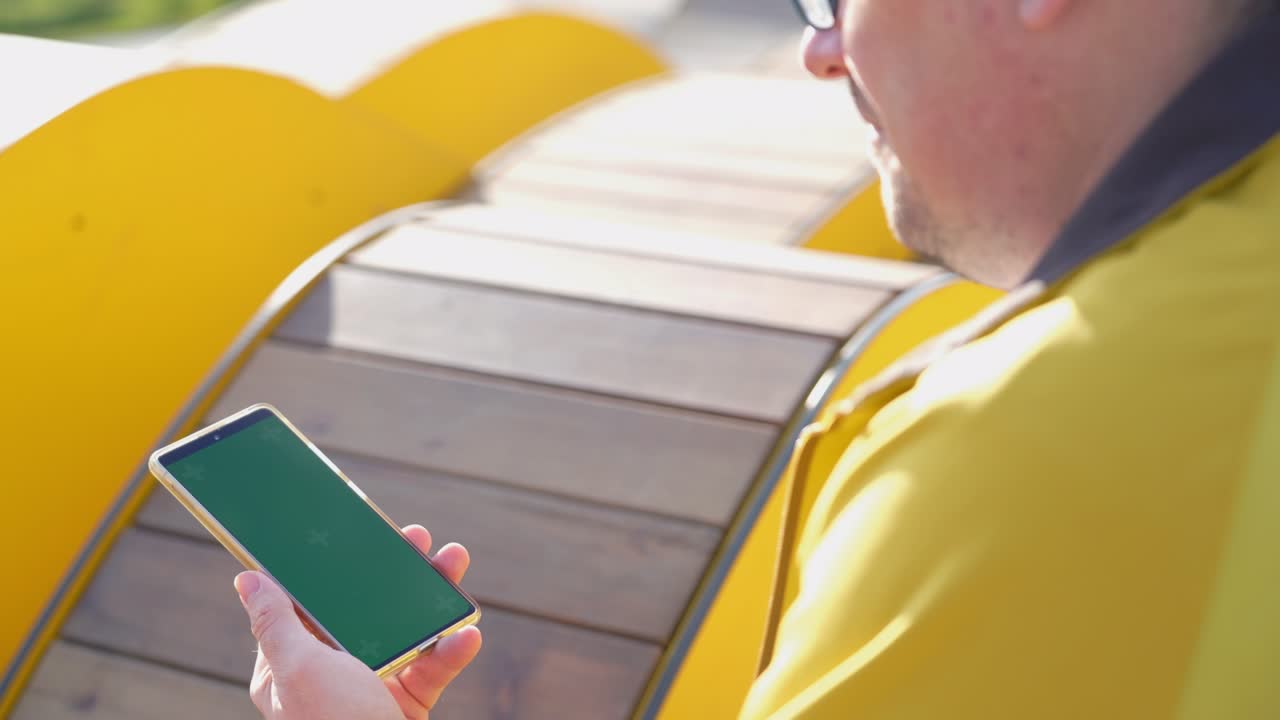 vista trasera de un hombre mirando un teléfono inteligente con pantalla verde y navegando en línea afuera. maquillaje para ver contenido. teléfono digital en blanco en la mano de un tipo sentado al aire libre en la calle del banco