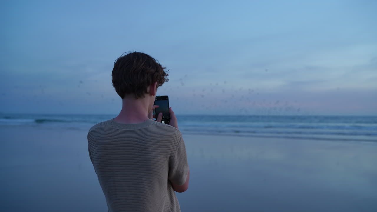 Teenager capturing picturesque moment, standing on wet sandy beach while documenting flying seabird flock against twilight ocean backdrop during golden hour photography session