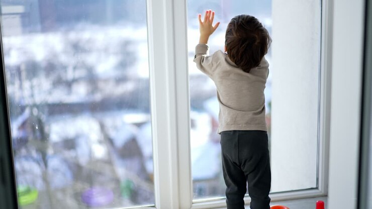 Rear view of a baby boy sitting at the panoramic window. Little child tap his hands by the window.