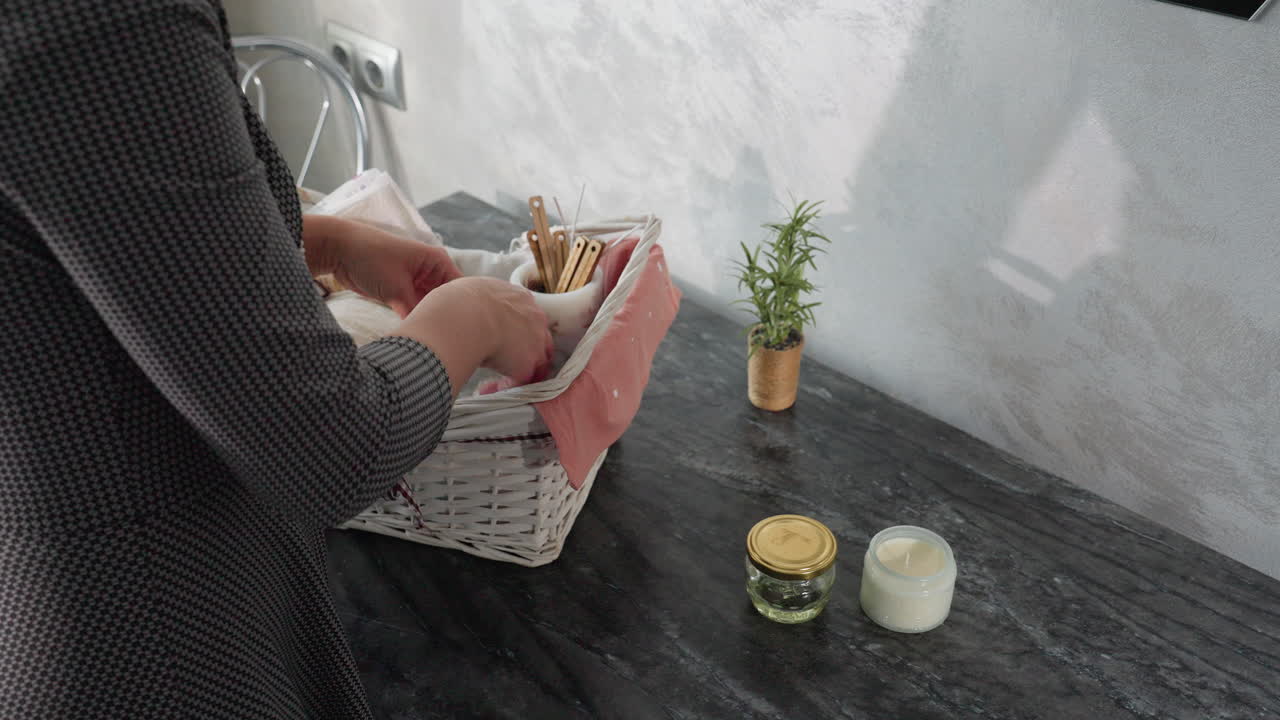 Partial side view of housewife in checkered dress unpacking basket and placing items like glass jar and candle on marble kitchen table near small potted plant under soft daylight in cozy modern home