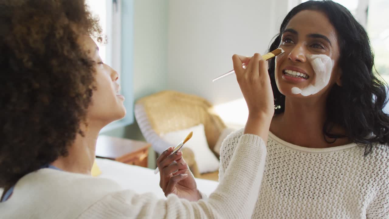 felices amigas diversas sentadas en la cama y aplicando una máscara facial de belleza