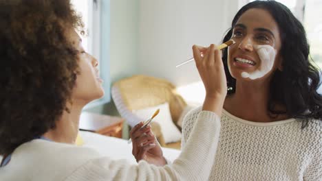 Happy-diverse-female-friends-sitting-on-bed-and-applying-beauty-face-mask