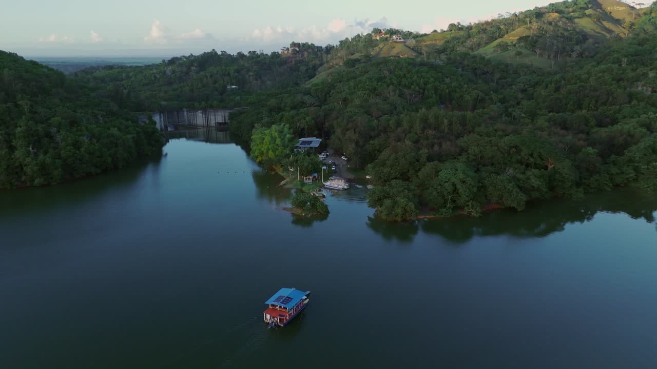 Ferry cruising through calm waters of Hatillo Dam reservoir, surrounded by lush green hills and peaceful landscape, perfect for travel or nature footage, Dominican Republic. Aerial backward at sunset