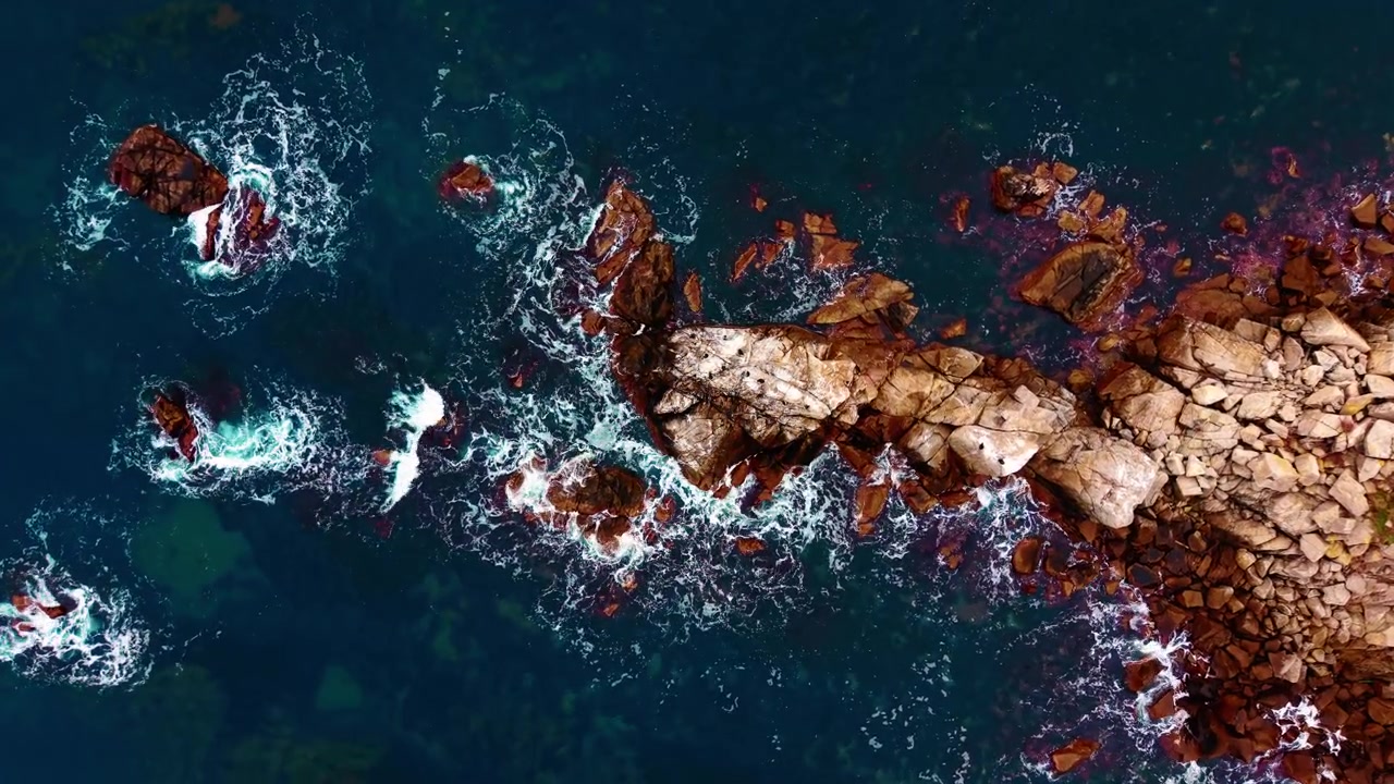 Aerial close up of waves on coastal rocks. Overhead shot captures white sea foam swirling around a narrow reddish rock point.